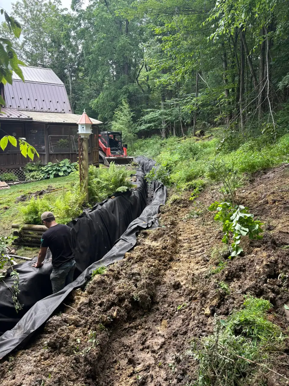 Red machinery and dark fabric piping near wooden structures and trees with exposed earth