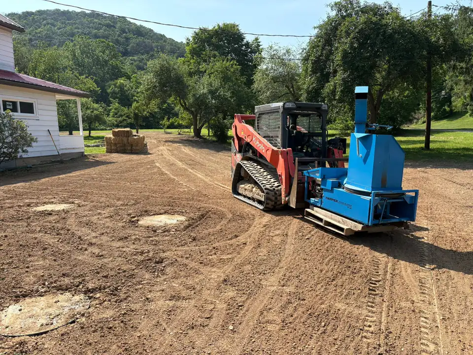 Red tracked machine and blue portable chipper on dirt ground near house with trees.
