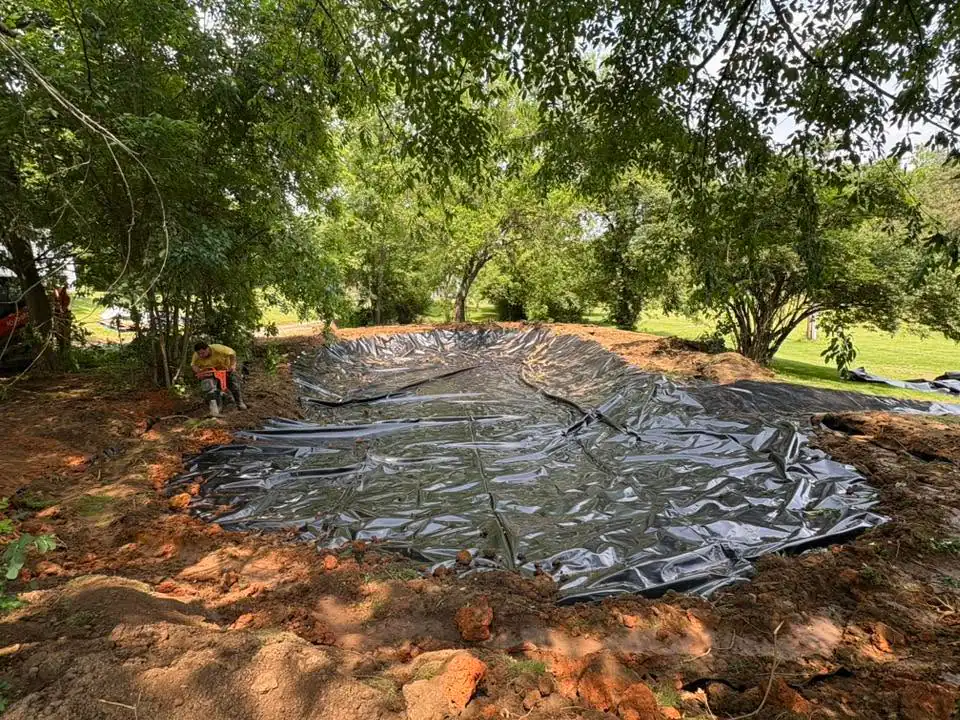 Black landscape fabric covering soil under mature trees in residential yard.