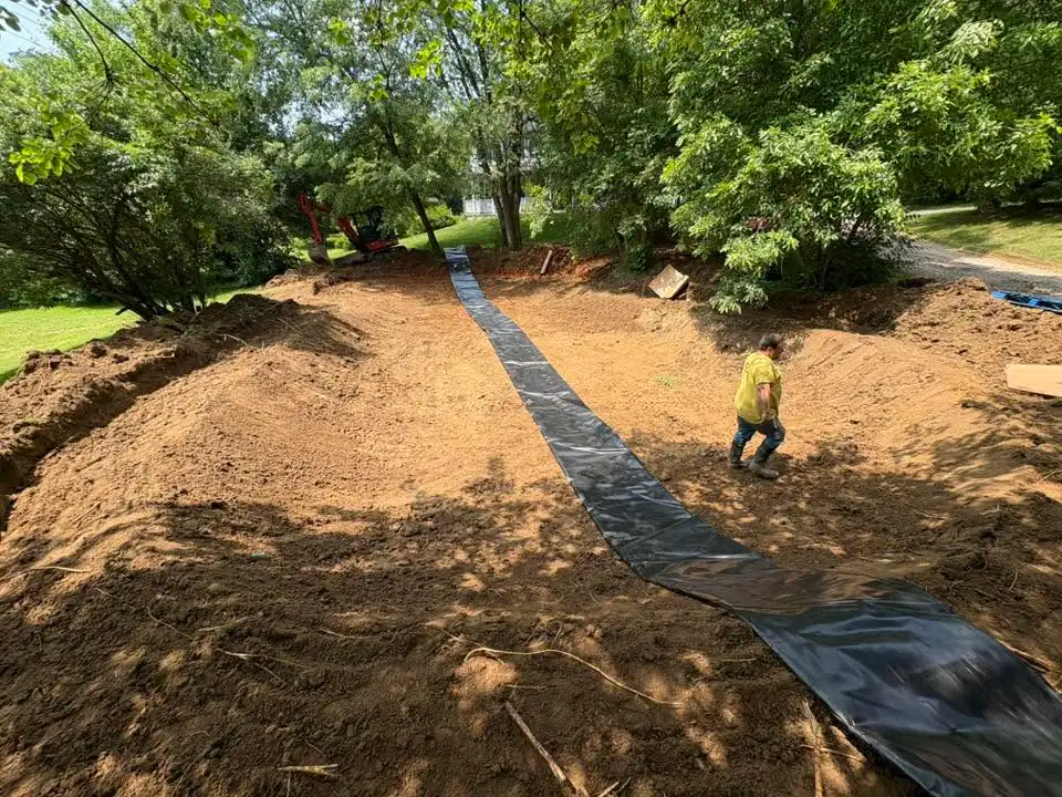 Person installing black landscape fabric on soil in residential yard under tree shade.