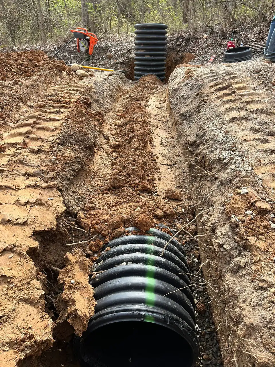 Black drainage pipe in trench with stacked pipes and worker in background