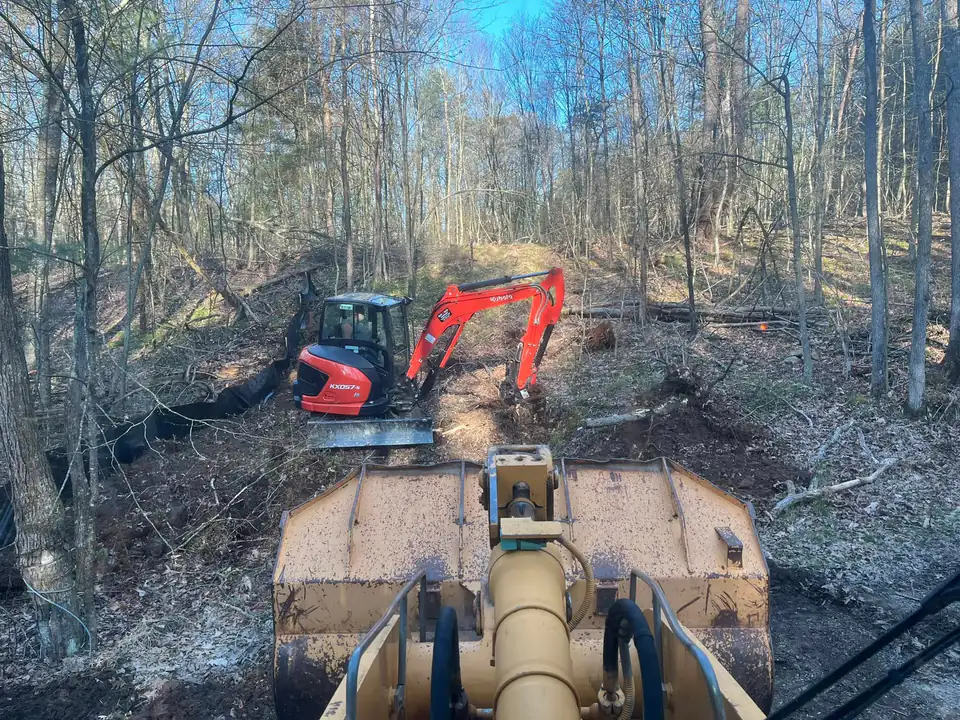 Operator's view over a loader bucket toward a red mini excavator in a wooded clearing