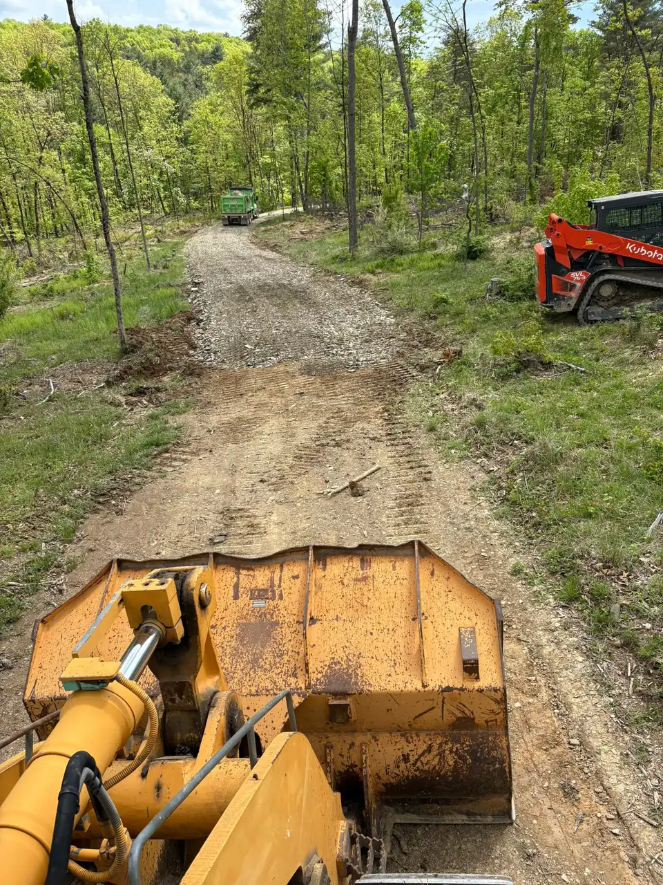 Operator's view over a loader bucket down a new gravel lane through the woods