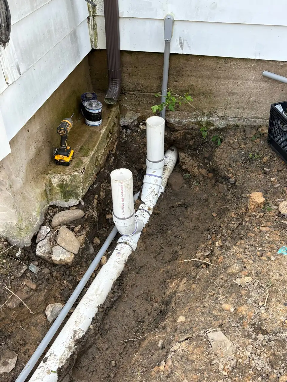 Excavated trench along a house foundation with white downspout drain pipes