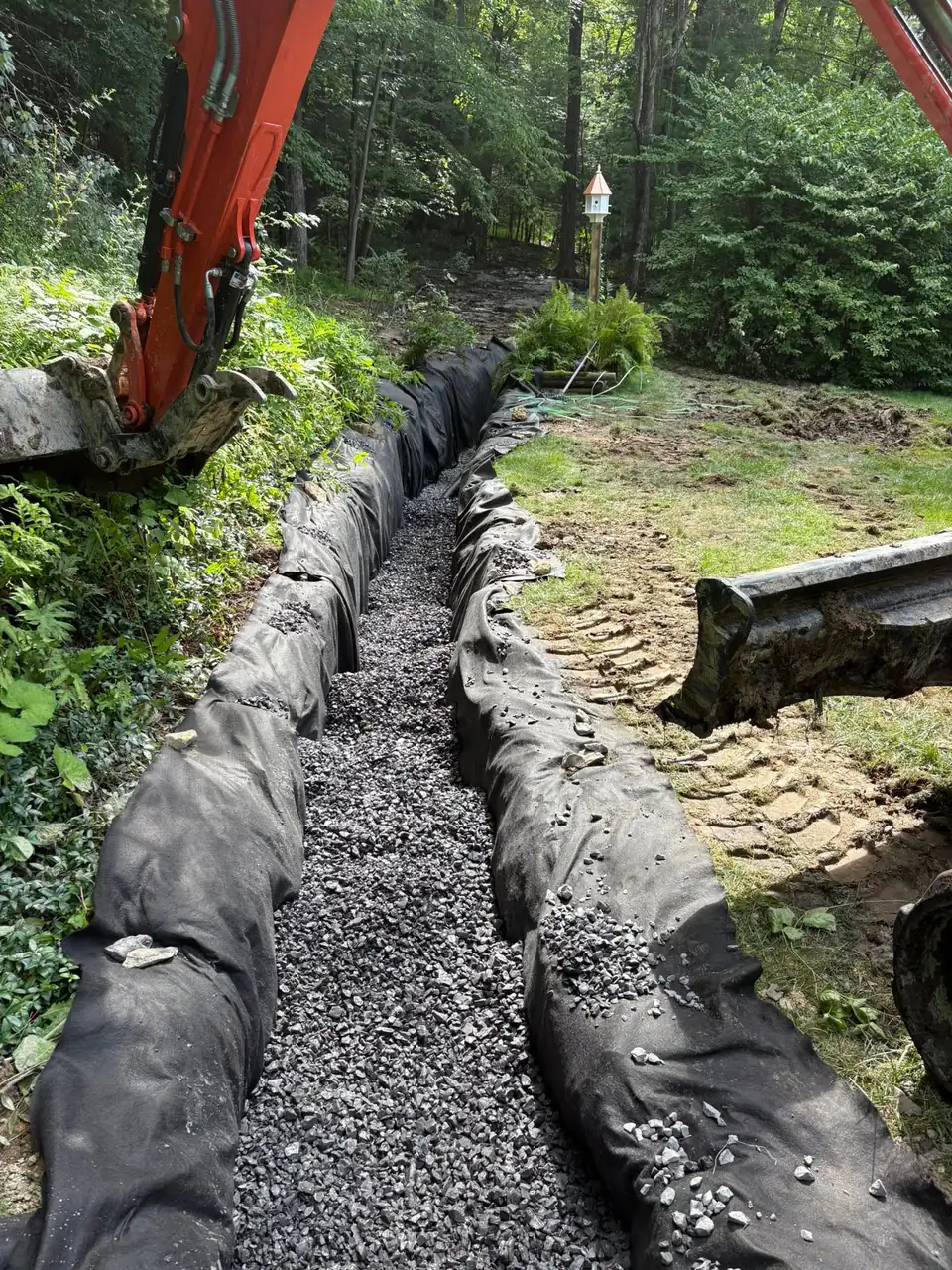 Sections of black corrugated pipe in prepared trench with gravel and red machinery.