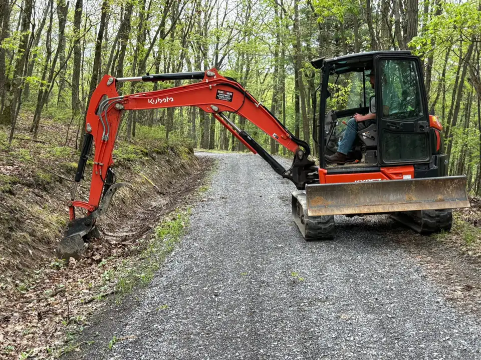 Red excavator on gravel road in wooded area with operator inside.