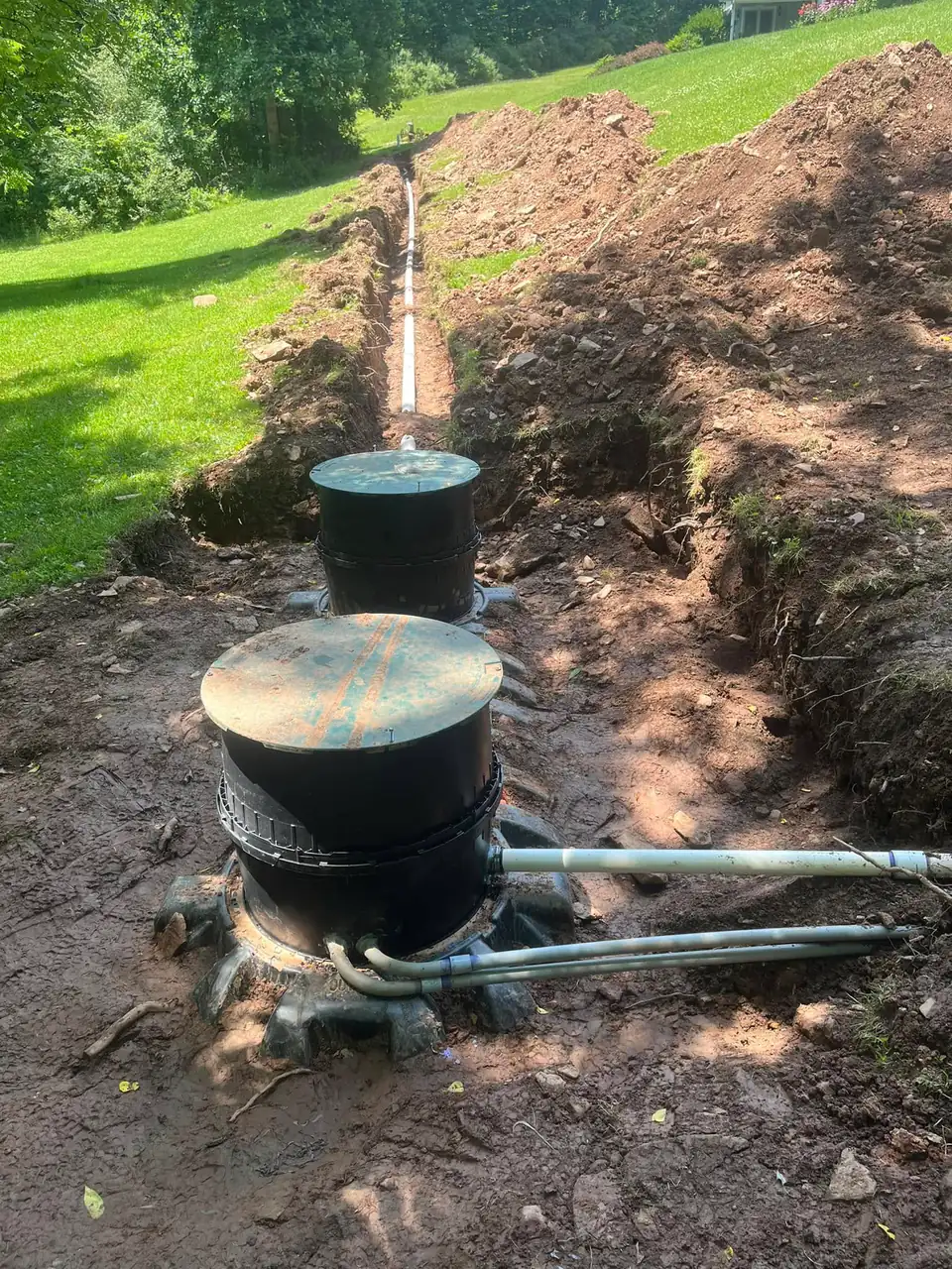 Black cylindrical septic tanks stacked in dirt trench with metal piping on grassy hillside.