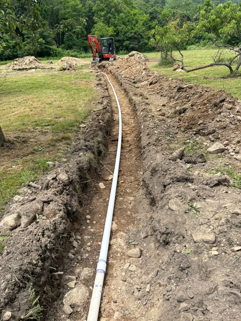 Trench with pipe across grassy field with excavator