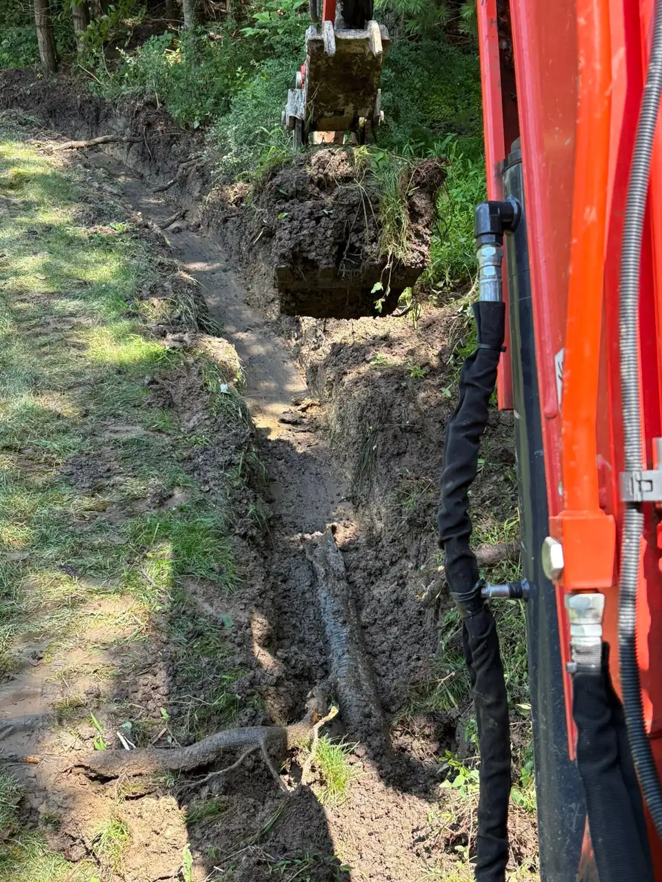 Excavator arm beside a narrow trench along a grassy bank