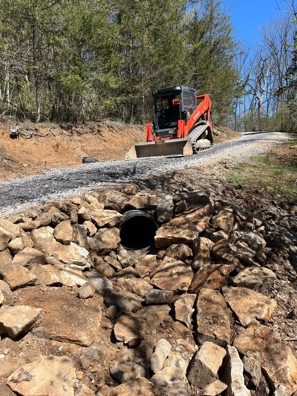 Tracked loader on a gravel drive above a culvert pipe set in a stacked-rock headwall