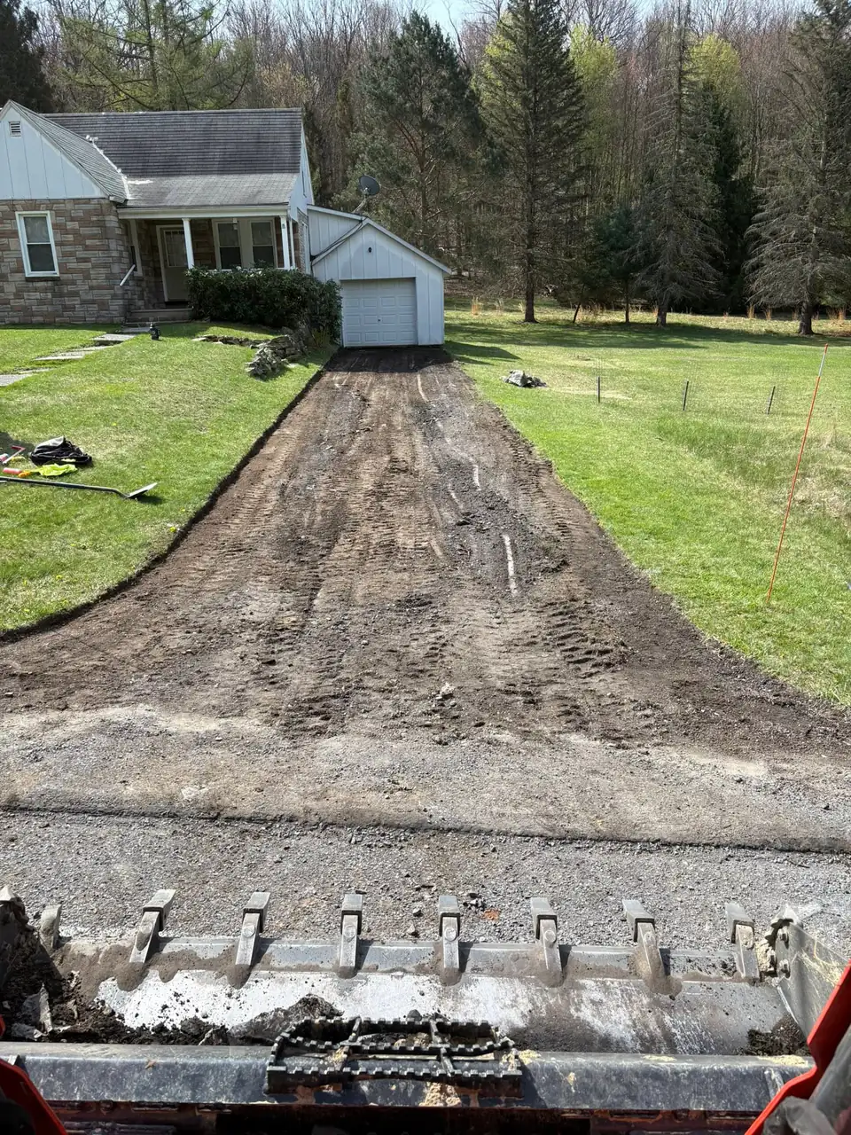 Graded driveway base leading to a stone house and garage, seen over a loader bucket