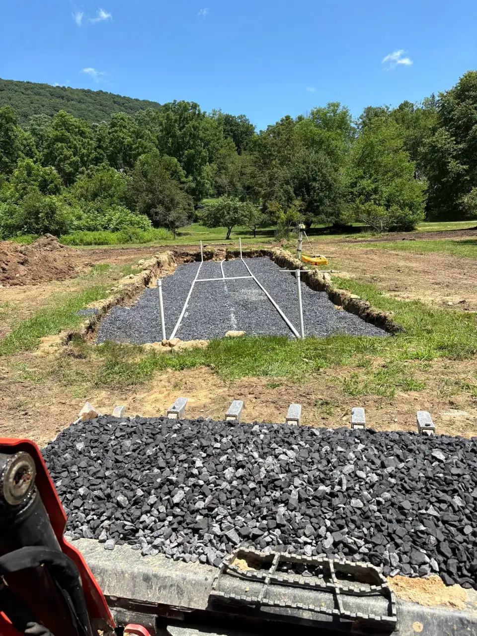 Operator's view over a bucket of stone toward a gravel bed with pipes in a grassy field